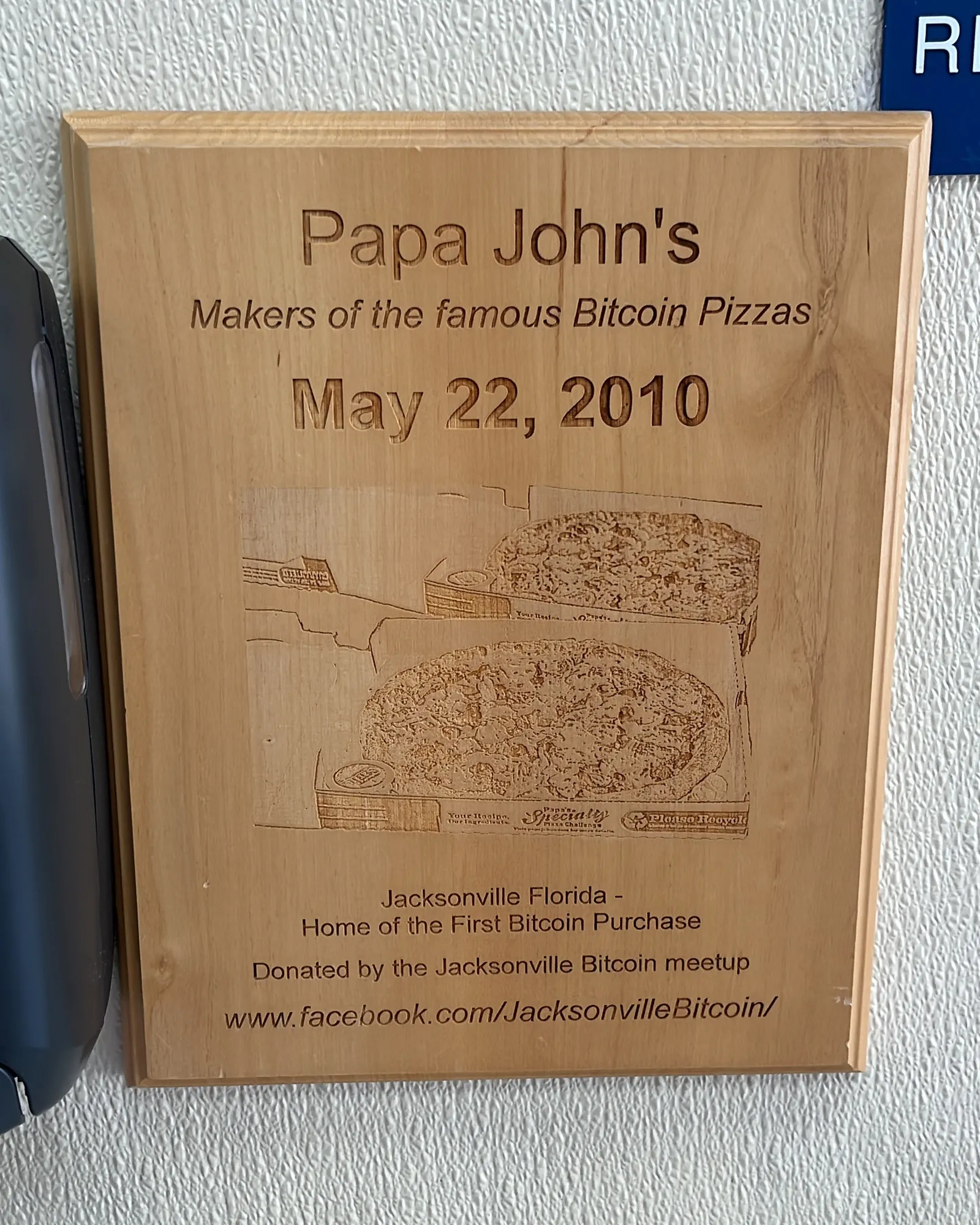 Una placa de madera en la pared con dos pizzas grabadas y el texto «Papa John’s. Makers of the famous Bitcoin Pizzas. May 22, 2010».
