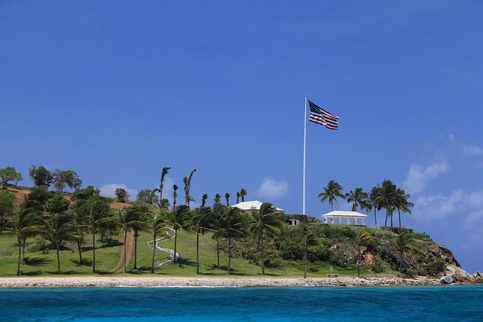 Uma ilha pequena com palmeiras e uma encosta gramada, no alto um mastro com a bandeira americana e um quiosque branco, água azul-turquesa em volta.