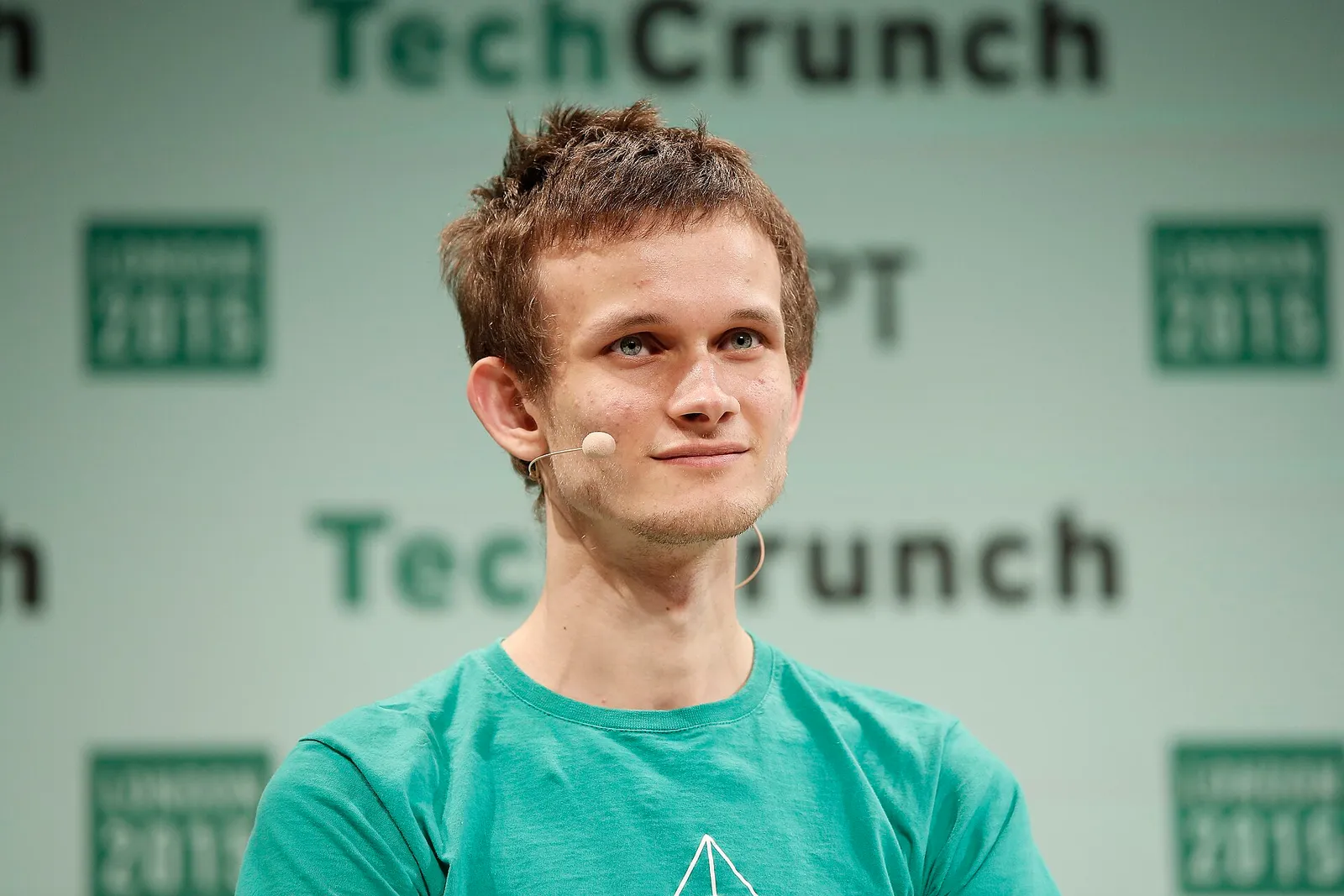 Un joven delgado con camiseta verde y un micrófono de diadema junto a la mejilla, delante de un panel de TechCrunch.